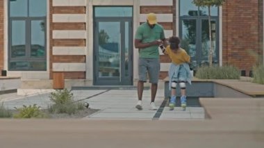 Siyahi babanın kameraya doğru yürüdüğü ve küçük kızı paten kaymayı öğrenirken desteklediği bir kare.