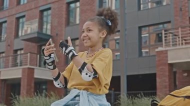 Koruyucu bileklik ve dirseklik takmış küçük Afrikalı Amerikalı kız sokakta paten kayıyor, kamerada gülümsüyor ve konuşuyor video ararken ya da video bloğu yaparken