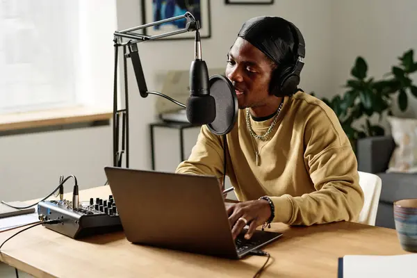 stock image Young African American man in headphones speaking or singing in microphone while sitting in front of laptop and typing on keyboard