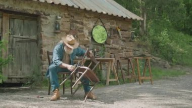 Ormandaki evin dışında sandalyeyi tamir ederken pense kullanan şapkalı yaşlı bir adamın uzun bir resmi.