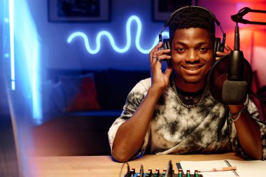 Kulaklıklı gülümseyen Afrikalı Amerikalı genç adam iş yerinde mikrofonun önünde oturmuş podcast yaparken kameraya bakıyor.