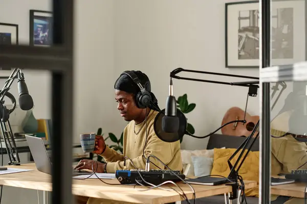 Young African American man with cup of coffee using laptop at break while preparing for recording new podcast with guest in studio