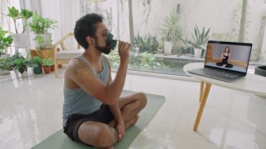 Spor kıyafetli bir adam evde egzersiz minderinde oturup su içiyor ve internetten kadın antrenörle konuşuyor.