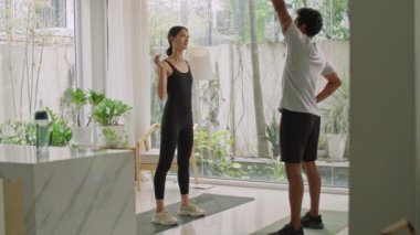 Spor kıyafetli genç, zayıf bir kadının evde antrenörle antrenman yaparken yan eğilmesiyle dolu bir fotoğraf.