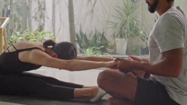 Genç bir kadın evde egzersiz yaparken kişisel fitness eğitmeninin yardımıyla öne doğru kıvrılıyor.