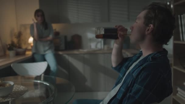 キッチンでテーブルに座ったボトルからアルコールを飲んで夫と口論する動揺する女性の中長いショット — ストック動画