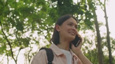 Güneşli yaz gününde parkta yürüyen ve cep telefonuyla konuşan genç bir kadının düşük açılı görüntüsü.