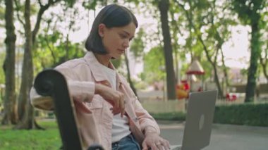 Yaz günü halka açık parkta bankta oturan ve internette dizüstü bilgisayarını karıştıran günlük kıyafetli genç bir kadın.