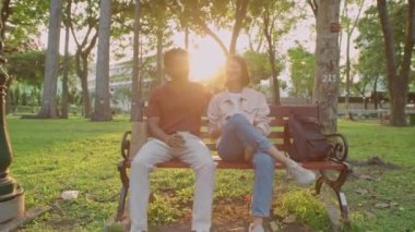 Gün batımında parkta bankta otururken kahve içmek ve konuşmak için bekleyen genç çiftin tam kavisli görüntüsü.