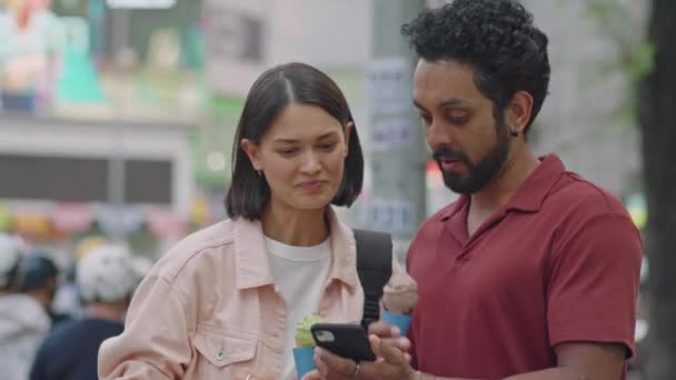 若いカップルのミディアムショット 携帯電話の都市マップを見て 路上で立ってアイスクリームを食べながらルートを議論 — ストック動画