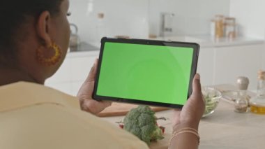 Siyah kadının elinde krom anahtar yeşil ekranlı dijital tablet tutarken evdeki mutfak masasında yemek pişirmeye hazırlandığı fotoğraf.