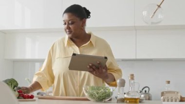 Orta boy Afro-Amerikan kadını dijital tablet üzerinde yemek tarifi okurken ve evde yemek pişirmek için yemek hazırlarken.