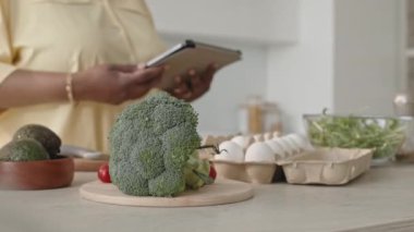 Mutfak masasının üzerinde taze brokoli ve diğer yemek malzemeleriyle yakın plan seçmeli fotoğraf, arka planda dijital tablet kullanan kadın.