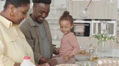 Küçük tatlı kız mutfak masasında oturuyor, anne ve babama salata pişirmede yardım ediyor ve evde birlikte yemek hazırlarken onlara beşlik çakıyor.