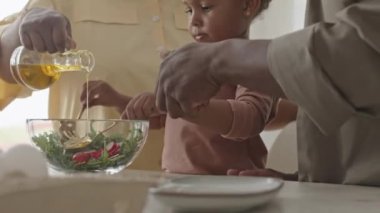 Afrika kökenli Amerikalı küçük bir kız, babasının yardımıyla cam kasede sebze salatasını karıştırırken anne zeytinyağı ekliyor.