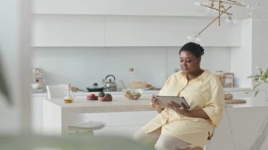 Afro-Amerikalı kadın mutfak masasında oturmuş evde yemek pişirmeden önce dijital tablette yemek tarifi arıyor.