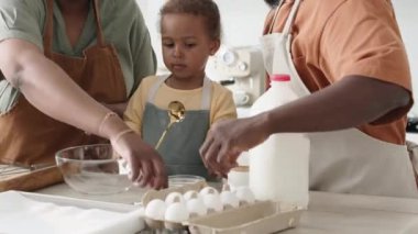 Afro-Amerikan ebeveynler ve tatlı küçük kız mutfak masasında birlikte yemek pişiriyorlar, anne yumurta kırıyor ve baba kızla konuşuyor.