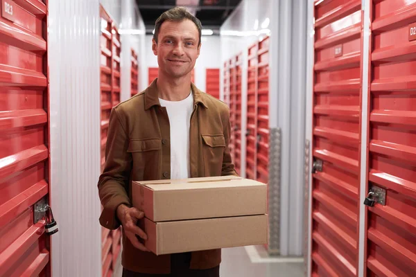 Smiling mature man with cardboard box standing in self-storage where he rents unit
