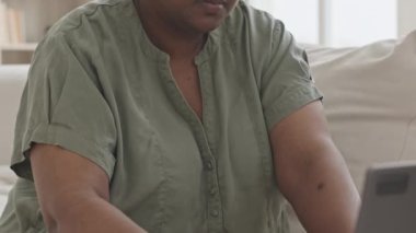 Siyahi bir kadının evdeki kanepede oturduğu, bilgisayarında çalıştığı ve dokümanları okuduğu bir fotoğraf.