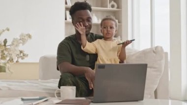 Askeri üniformalı Afro-Amerikan askeri oturma odasındaki kanepede dizüstü bilgisayarla oturuyor ve tatlı küçük kızıyla oynuyor.