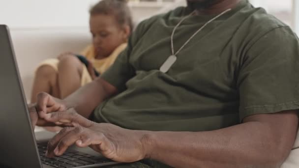 一个身穿军服的非裔美国人坐在家里的沙发上 在笔记本电脑上浏览互联网 而他的小女儿则坐在他旁边 一边打电话一边拍着照片 — 图库视频影像