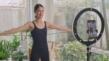 Kadın fitness blogcusu, evde sosyal medya kanalı için video çalışması kaydedilirken tripodlu telefonun önünde direnç bandıyla egzersiz yapıyor