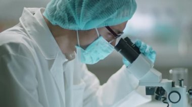 Koruyucu yüz maskesi, şapka ve eldiven takmış, mikroskoptan bakan ve laboratuarda çalışırken notlar alan Asyalı erkek bilim adamı.