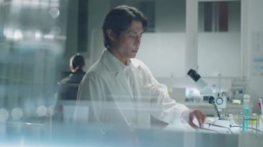 Laboratuar önlüklü Asyalı bilim adamının cam duvarından iş yerine yürürken, gözlük takarken, mikroskop kullanırken ve laboratuvar araştırmaları sırasında not alırken görüntüsünü al.
