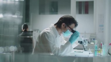 İş yerinde oturan, mikroskop kullanan ve laboratuvarda çalışırken araştırma sonuçlarını yazan koruyucu maske takan Asyalı bilim adamlarının yan görüntüsü