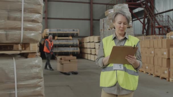 彼女の男性の同僚が倉庫の段ボール箱が付いているパレットを再配置している間 報告書を満たす女性のマネージャーの中間の長いショット — ストック動画