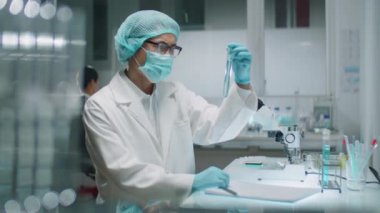 Erkek kimyagerin koruyucu yüz maskesi, tıbbi şapka ve eldiven taktığı test tüpünde sıvı maddeyi incelerken ve laboratuarda çalışırken notlar yazarken orta boy fotoğrafı.
