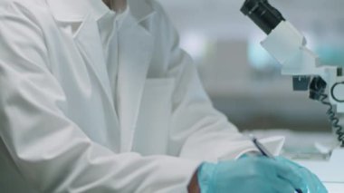 Laboratuvar önlüklü ve eldivenli Asyalı bilim adamının laboratuvar masasında çalışırken not alıp tüp yuttuğu görüntüleri.