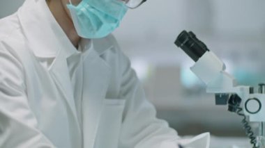 Asyalı bilim adamının koruyucu maskeli, tıbbi şapkalı ve eldivenli görüntüsü laboratuarda yapılan deneyin sonuçlarını yazıyor.