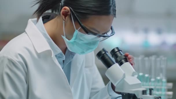 在实验室工作时 使用显微镜和安全眼镜拍摄戴防护面罩和安全眼镜的亚洲女科学家的照片 — 图库视频影像
