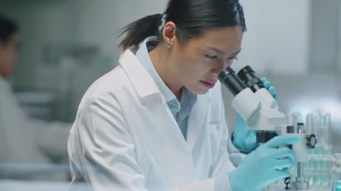 Beyaz önlüklü ve koruyucu eldivenli kadın Asyalı bilim adamının laboratuvar araştırmaları sırasında mikroskop kullanırken orta boy fotoğrafı.