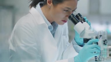 Laboratuvarda araştırma yaparken, laboratuvar önlüklü ve steril eldivenli kadın Asyalı bilim adamının mikroskoptan bakarken görüntüsünü yukarı kaldır.