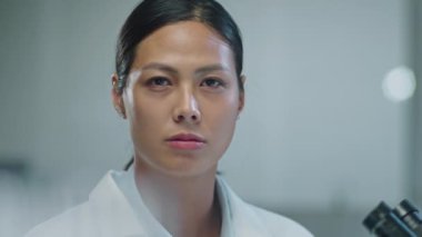 Beyaz önlüklü ciddi bir Asyalı kadın bilim adamının portresi laboratuvarda çalışırken kameraya özgüvenle bakıyor.