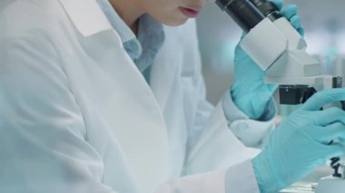 Laboratuvar önlüklü ve tıbbi eldivenli kadın Asyalı bilim adamının bileşik mikroskop kullanarak ve laboratuvarda araştırma yaparken notlar yazarken aşağı eğilmesini.
