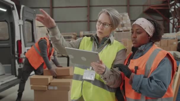 バックグラウンドでボックスをバンにロードしながら貯蔵の再配置の計画を議論する倉庫労働者の多文化カップルの中型ショット — ストック動画