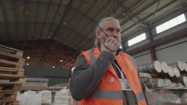 Depoda çalışırken yansıma giysileri giyen ve telsizle konuşan olgun bir adamın düşük açılı portresi.