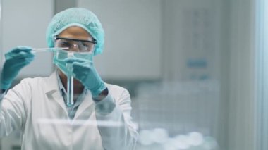 Korumacı yüz maskesi, tıbbi şapka ve eldivenli Asyalı kadın bilim adamının laboratuvar deneyi sırasında deney tüpüne pipetle mavi kimyasalı dökerken orta boy fotoğrafı.