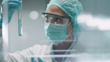 Koruyucu maskeli, şapkalı ve eldivenli Asyalı kadın bilim adamlarının cam duvarından, deney tüpüne pipetli mavi kimyasalları dökerek laboratuvar deneylerindeki tepkilere bakın.