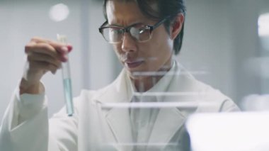 Laboratuvarda cam duvarın arkasında duran Asyalı kimyagerin, deney tüpüne pipetle mavi kimyasalı dökerek, karıştırarak ve deney sırasında reaksiyonu izleyerek,