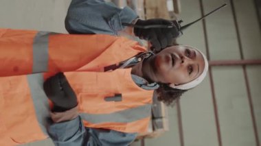 Afro-Amerikan lojistik operatörünün dikey portresi depoda telsiz kullanıyor ve kameraya bakıyor.