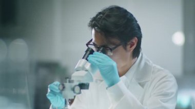 Medikal eldivenli ve beyaz önlüklü erkek Asyalı bilim adamının mikroskopla çalışırken ve laboratuvar deneyleri sırasında notlar alırken orta boy fotoğrafı.