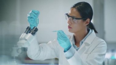 Beyaz önlüklü, koruyucu gözlüklü ve eldivenli Asyalı kadın kimyagerin mavi kimyasallarla deney yaparken pipet ve test tüpü kullanırken orta boy fotoğrafı.