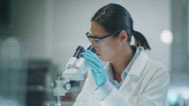 Medikal eldivenli, koruyucu gözlüklü ve beyaz önlüklü Asyalı kadın bilim adamının mikroskoba bakarken ve laboratuvar deneyleri sırasında notlar alırken orta boy fotoğrafı.