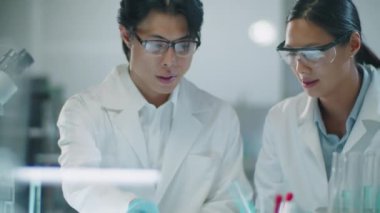 Korumacı eldivenler ve gözlükler takmış Asyalı erkek ve kadın bilim adamlarının petri kabına pipetle kimyasallar dökerek laboratuvardaki deneyleri tartışırkenki görüntüleri.