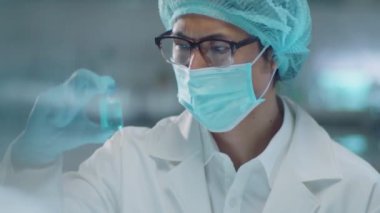 Korumacı maskeli, tıbbi şapkalı ve eldivenli Asyalı erkek kimyagerin laboratuvarda deney yaparken mor kimyasalla şişeye baktığı görüntüyü yukarı kaldır.