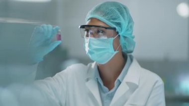 Korunaklı yüz maskesi takan Asyalı kadın kimyager, tıbbi şapka ve eldivenler laboratuvar araştırmaları sırasında kimyasalla şişeyi inceliyor.